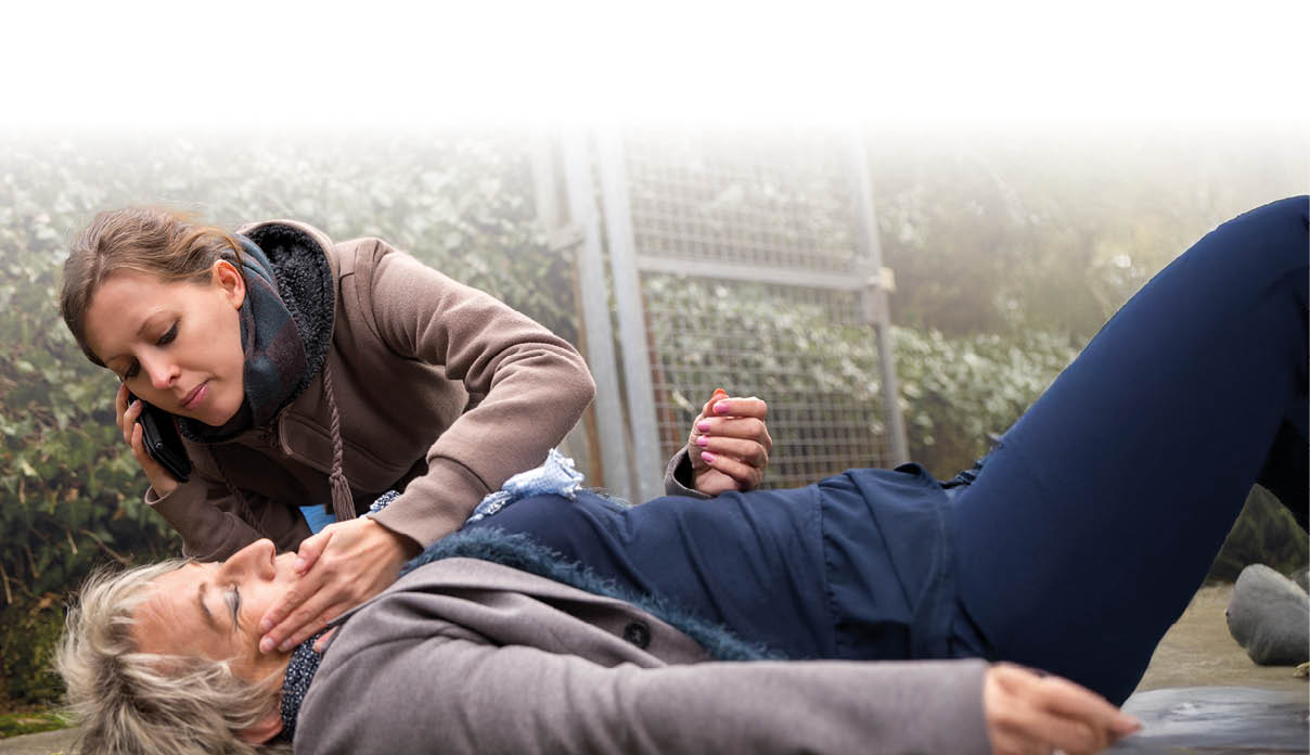 Eine ltere Frau liegt bewusstlos auf dem Boden. EIne j ngere Frau kniet neben ihr und beugt sich  ber sie. Sie ber hrt ihr Gesicht und versucht sie anzusprechen.