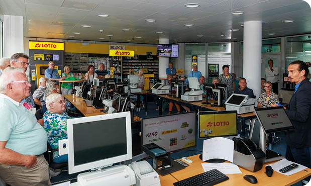 Auf dem Bild ist ein Schulungs- oder Informationsraum von LOTTO Rheinland-Pfalz zu sehen. Mehrere Personen sitzen oder stehen zwischen Verkaufstheken und Kassenarbeitspl tzen, die mit Monitoren, Druckern und Terminals ausgestattet sind.   Im Vordergrund steht ein Mann im Anzug, der offenbar einen Vortrag h lt oder eine Einweisung gibt. Die Anwesenden –  berwiegend  ltere Personen – h ren aufmerksam zu. An den Theken sind Logos und Hinweise zu LOTTO, KENO und weiteren Produkten angebracht.    Die Umgebung  hnelt einem realit tsnah eingerichteten Verkaufsraum, wie er in einer Lotto-Annahmestelle zu finden ist.