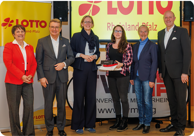 Auf dem Bild ist eine Gruppe von sechs Personen zu sehen, die nebeneinander vor einer gelben Pr sentationswand mit dem Schriftzug LOTTO Rheinland-Pfalz stehen.   In der Mitte h lt eine junge Frau gemeinsam mit einer weiteren Person eine kleine Auszeichnung beziehungsweise ein Pr sent in den H nden. Die  brigen Personen stehen l chelnd daneben, offenbar im Rahmen einer offiziellen  bergabe oder Ehrung.    Alle Beteiligten sind gepflegt gekleidet – teils im Business-Outfit mit Anzug oder Blazer.