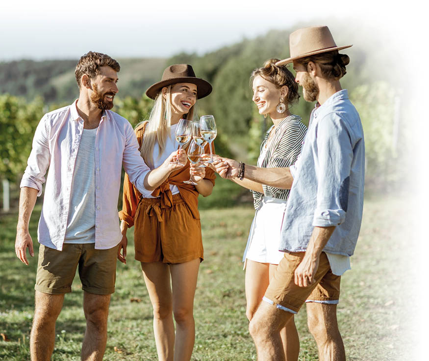 Das Bild zeigt vier junge Erwachsene, die in einem Weinberg stehen und gemeinsam mit Weingl sern ansto en. Zwei M nner und zwei Frauen stehen im Halbkreis auf einer Wiese zwischen Rebreihen. Im Hintergrund sind gr ne Weinreben und sanfte H gel unter einem klaren Himmel zu sehen.   Alle vier l cheln und wirken entspannt und fr hlich. Sie halten jeweils ein Glas mit hellem Wei wein in der Hand und f hren die Gl ser in der Mitte zusammen. Die Personen tragen sommerliche Kleidung: leichte Hemden, Shorts, eine Frau tr gt einen braunen Hut und einen lockeren, braunen Zweiteiler, die andere eine gestreifte Bluse und einen hellen Rock. Auch einer der M nner tr gt einen Hut. Das warme Sonnenlicht beleuchtet die Szene und erzeugt eine freundliche, gesellige Atmosph re an einem sonnigen Tag im Freien.