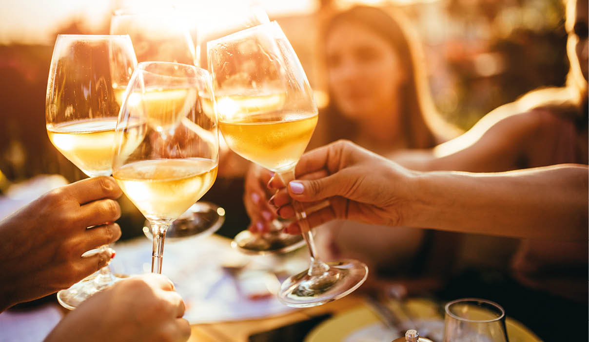 Das Bild zeigt eine Nahaufnahme von vier Weingl sern, die in der Mitte zusammengesto en werden. Die Gl ser sind mit hellem Wei wein gef llt, der im warmen Licht golden schimmert. Mehrere H nde halten die Gl ser am Stiel fest; die Personen selbst sind nur teilweise oder unscharf im Hintergrund zu erkennen.   Das Sonnenlicht scheint direkt von hinten in die Szene und erzeugt starke, warme Orange- und Goldt ne. Die Gl ser reflektieren das Licht, sodass helle Lichtpunkte und Spiegelungen sichtbar sind. Im Hintergrund ist eine Person unscharf zu sehen, vermutlich an einem Tisch im Freien sitzend. Unten im Bildrand sind Teile eines gedeckten Tisches erkennbar, darunter ein weiteres Glas und Geschirr. Die Stimmung wirkt gesellig und feierlich, vermutlich bei einem Treffen im Freien w hrend des Sonnenuntergangs.