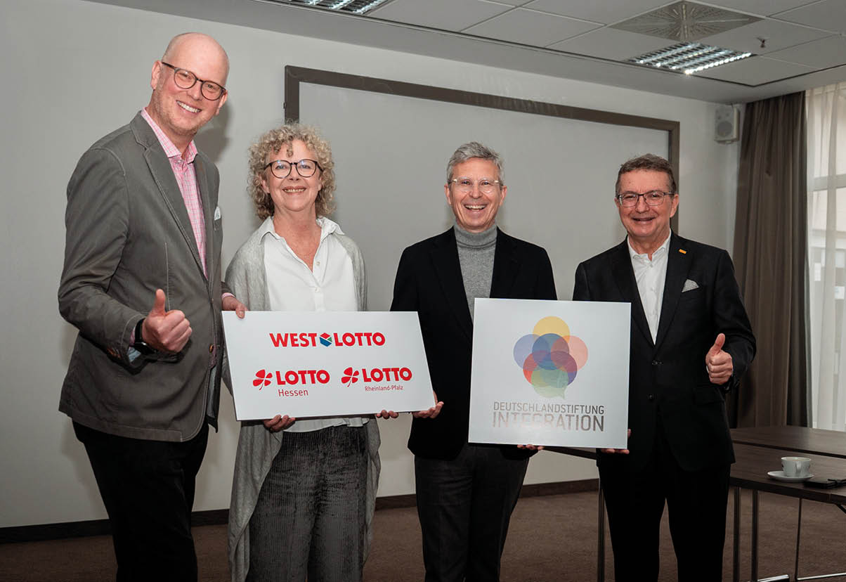 Vier Personen, eine Frau und drei M nner, halten zwei Tafeln hoch. Auf der einen stehen die Logos der drei Lotto-Gesellschaften Westlotto, Lotto Hessen sowie Lotto Rheinland-Pfalz. Auf der anderen steht das Logo der Deutschlandstiftung Integration. Alle strahlen zufrieden in die Kamera. Die beiden M nner rechts und links halten den Daumen hoch.
