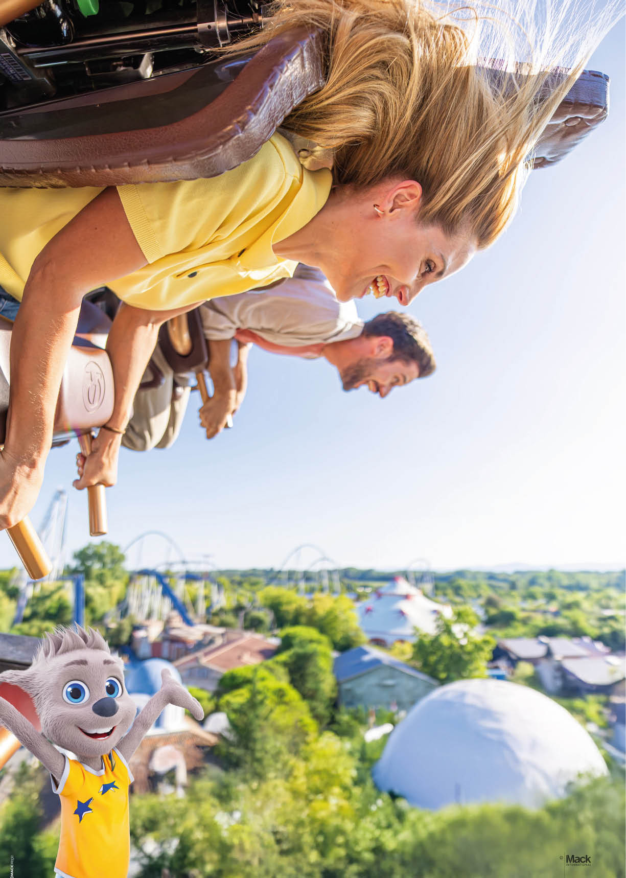 Oben im Bild, im Vordergrund:   Zwei Personen sitzen nebeneinander in einer Wagenreihe eines Freizeitpark-Fahrgesch fts. Sie sind leicht nach vorn geneigt, so als w rde die Fahrt gerade eine Abw rtsbewegung oder einen schnellen Schlenker machen.  Die Person links tr gt ein hellgelbes, kurz rmeliges Poloshirt. Ihr langes, hellblondes Haar weht nach hinten. Beide H nde greifen an goldfarbene Haltegriffe an der Vorderkante des Sitzes.  Die Person rechts tr gt ein helles Oberteil und h lt ebenfalls beide H nde an einem Haltegriff. Die Gesichter der beiden Personen sind in dem Bild teilweise unscharf bzw. aus Datenschutzgr nden weichgezeichnet; feinere Mimik ist nicht erkennbar.  Mittel- und Hintergrund:    Unterhalb der Fahrg ste breitet sich ein Freizeitpark mit vielen B umen, Geb uden und Fahrattraktionen aus.  In der Ferne sind mehrere Achterbahnstrecken mit geschwungenen Schienen gut sichtbar; sie ziehen sich  ber den Horizont.  Rechts unten im Bild steht eine gro e wei e Kuppel. Daneben sind weitere Geb ude und gr ne Baumkronen zu sehen.  Der Himmel ist klar und hellblau, das Licht wirkt sonnig und erzeugt helle, frische Farben.  Vordergrund, unten links:    Ein fr hliches, cartoonartiges Maskottchen in Form einer kleinen, hellgrauen Tierfigur ( hnlich einer Maus oder eines kleinen Wolfs) steht mit beiden Armen nach oben gestreckt. Es hat gro e blaue Augen, ein breites L cheln und tr gt ein gelbes  rmelloses Oberteil mit zwei blauen Sternen. Die Gestik wirkt einladend und jubelnd.  Farben und Stimmung:    Die Szene ist hell und sommerlich: viel Gelb (Kleidung, Sonnenlicht), viele Gr nt ne (B ume) und das helle Blau des Himmels.  Die Gesamtstimmung ist lebhaft und freudig, vermittelt Bewegung, Spa  und den Nervenkitzel einer Vergn gungspark-Fahrt.