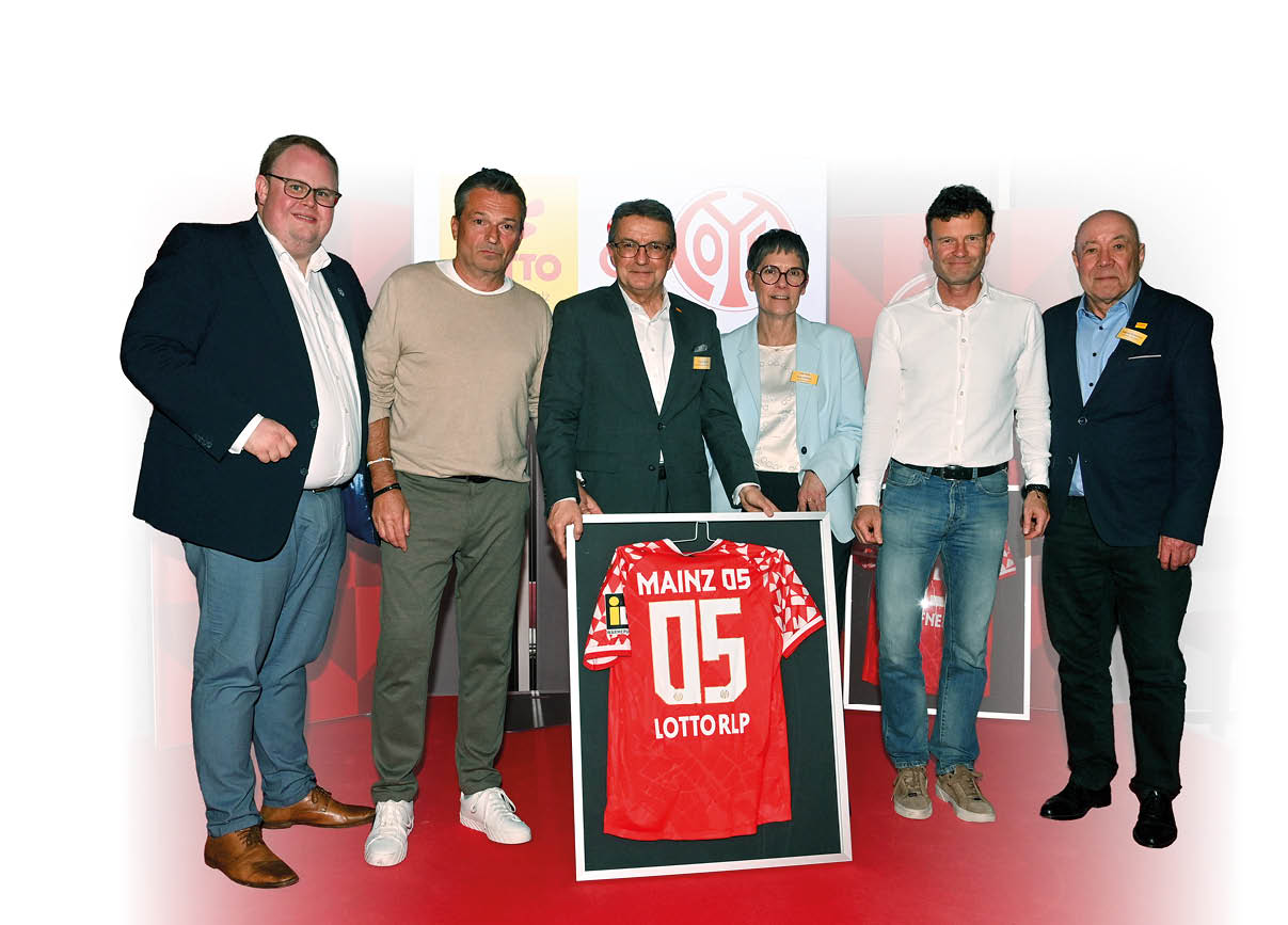 Auf dem Bild stehen sechs Erwachsene nebeneinander auf einer B hne und pr sentieren gemeinsam ein gerahmtes Fu balltrikot. Der Hintergrund ist hell mit roten grafischen Elementen und Logos, die auf den Fu ballverein Mainz 05 hinweisen.   In der Mitte der Gruppe halten zwei Personen ein gro es, schwarz gerahmtes Trikot hoch. Das Trikot ist rot mit wei en Details und tr gt die Aufschrift „Mainz 05“ sowie die R ckennummer 05. Darunter steht der Schriftzug „LOTTO RLP“. Das Trikot wirkt wie ein Pr sent oder eine Ehrung und ist professionell eingerahmt.    Die sechs Personen stehen dicht nebeneinander und schauen in Richtung Kamera.    Ganz links steht ein kr ftiger Mann in einem dunklen Sakko, hellem Hemd und Jeans.    Neben ihm ein Mann in einem beigen Pullover und gr ner Hose.    In der Mitte steht ein  lterer Mann im dunklen Anzug mit wei em Hemd, der das Trikot mit pr sentiert.    Daneben eine Person mit kurzer Frisur, Brille und hellem Blazer, ebenfalls mit einem kleinen gelben Namensschild.    Rechts davon ein gro er Mann im wei en Hemd und Jeans.    Ganz rechts steht ein  lterer Mann im dunklen Anzug mit gelbem Namensschild.    Alle wirken entspannt und leicht l chelnd, als w rden sie f r ein offizielles Pressefoto posieren. Die B hne ist rot beleuchtet, was farblich gut zum roten Fu balltrikot passt. Das Bild vermittelt den Eindruck einer feierlichen  bergabe oder Ehrung im Zusammenhang mit dem Fu ballverein Mainz 05. ⚽