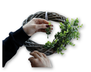 Eine Person legt gr ne Zweige in einen runden, geflochtenen Holzkranz auf einer hellen Arbeitsfl che. Hauptmotiv ist das Basteln eines Dekokranzes.