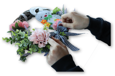 Ein runder, geflochtener Weidenkranz liegt frontal vor wei em Hintergrund. Links oben ist der Kranz mit gr nen, kleinen Bl ttern und einigen lila‑farbenen Bl tenrispen geschm ckt. Unten rechts ist eine graue Filz‑Hasenfigur befestigt, die seitlich am Kranz anliegt. Der Hase hat schwarze Punktaugen, ein rotes herzf rmiges N schen und h lt orange‑gelbe Filzblumen sowie eine gr ne Schleife. Das Bild zeigt eine fertige, fr hlingshafte oder sterliche T r‑ bzw. Tischdekoration.