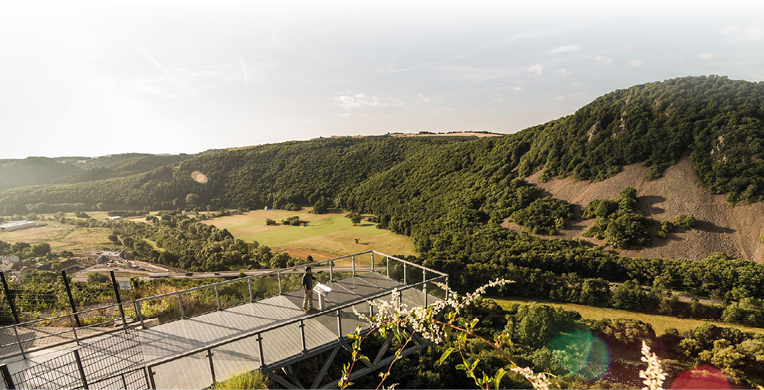 Auf dem Bild ist eine freitragende Aussichtsplattform aus Stahl zu sehen, die weit ber ein bewaldetes Tal hinausragt. Eine Person steht auf der Plattform und blickt auf die Landschaft. Unterhalb erstrecken sich Wiesen, Felder und ein Flusslauf, die H nge sind dicht bewaldet, teils mit steinigen Ger llfl chen.  Es handelt sich um den Nahe-Skywalk, eine markante Aussichtskonstruktion oberhalb des Nahetals bei Kirn.  