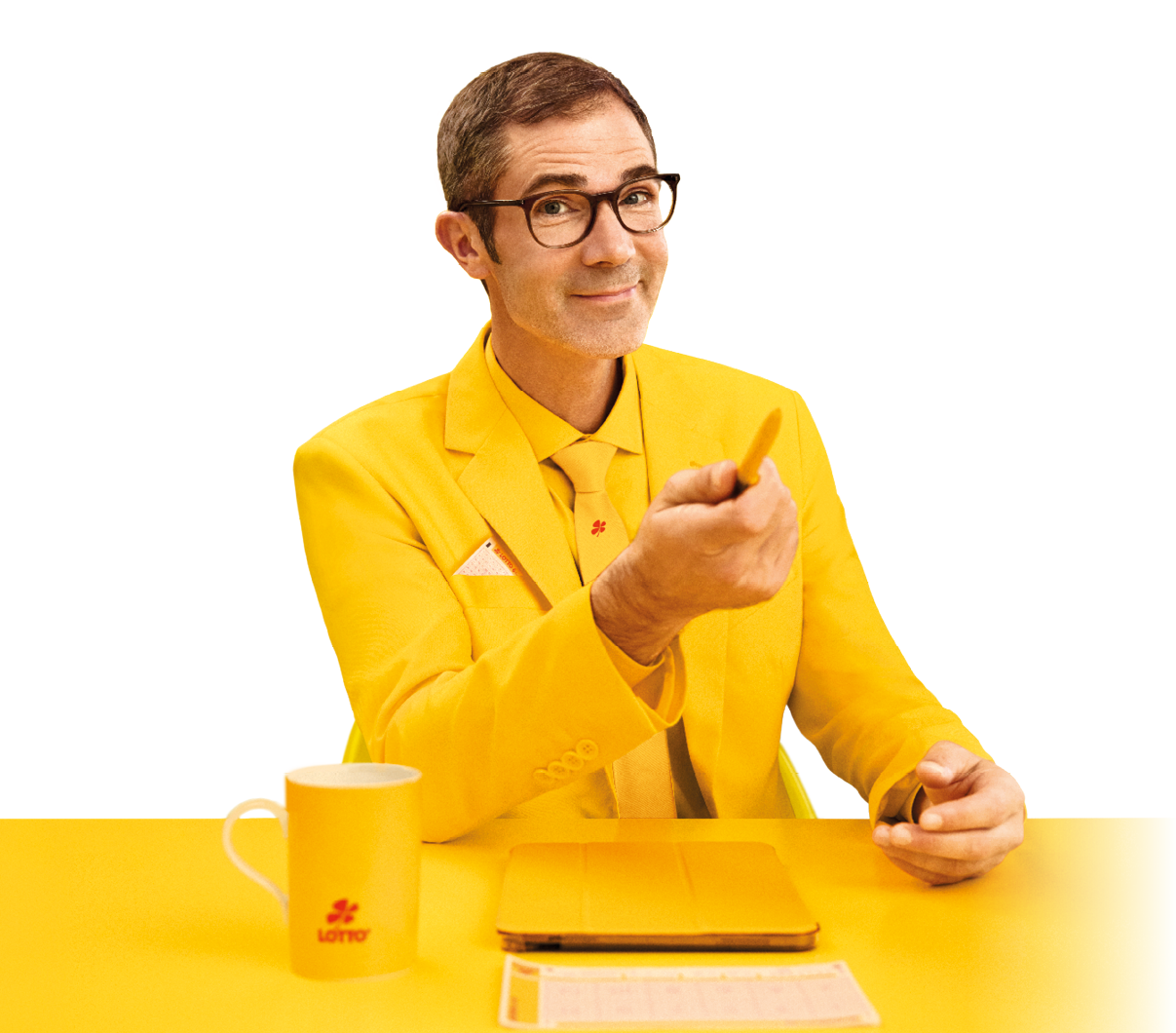 Ein Mann im gelben Anzug, gelben Hemd und Krawatte streckt einen Stift in Richtung Kamera. Vor ihm liegt ein Ipad in einer gelben Schutzh lle, ein Lottoschein auf einem gelben Tisch. Daneben steht ein eine gelbe Tasse mit dem Lotto-Logo. Er l chelt freundlich und wissend in die Kamera.