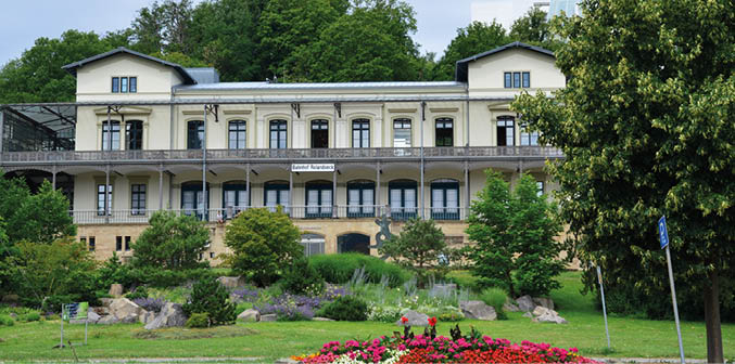 Ansicht eines historischen Bahnhofsgeb udes. Es handelt sich um das Museum Rolandseck. Vor dem hellen Geb ude erstreckt sich ein gepflegte Parklandschaft.