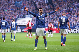 Ein Fu ballspieler in blau-wei er Mannschaftsuniform steht mittig mit weit ge ffneten Armen auf dem Spielfeld, vermutlich in einer Gestik des Feierns oder Appellierens. Mehrere weitere Spieler sind im Bildhintergrund verteilt; die Zuschauertrib ne ist voll und unscharf, sodass Stimmung und Menschenmenge erkennbar sind. Das Spielfeld ist gr ner Rasen, die Szene wirkt wie w hrend eines Ligaspiels im Stadion.