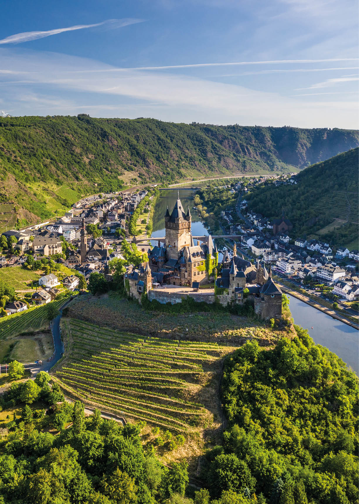Das Bild zeigt eine weite Landschaft mit Fluss, Ort und Burg aus erh hter Perspektive.   Auf der linken Seite thront auf einem steilen, dicht bewaldeten H gel eine mittelalterliche Burganlage mit mehreren T rmen und spitzen D chern. Der h chste Turm ragt deutlich  ber die anderen Geb ude hinaus. Die Burg wirkt massiv und aus dunklem Stein gebaut.    Unterhalb der Burg erstrecken sich Weinberge und gr ne H nge, die sich terrassenartig den H gel hinunterziehen.    Rechts im Bild flie t ein breiter, ruhiger Fluss durch das Tal. Entlang des Ufers liegt ein kleiner Ort mit dicht stehenden H usern, Stra en und einer Uferpromenade. Mehrere wei e Ausflugsschiffe sind am Fluss zu sehen.    Eine Br cke  berspannt den Fluss und verbindet beide Seiten des Tals.    Die umliegenden H gel sind gr n bewachsen und ziehen sich bis in die Ferne. Dar ber spannt sich ein hellblauer Himmel mit d nnen, langgezogenen Wolken.