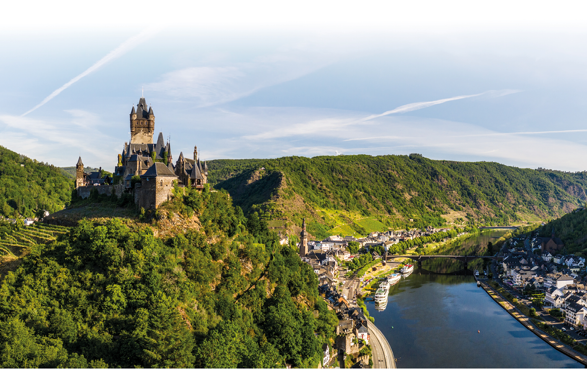 Das Bild zeigt eine weite Landschaft mit Fluss, Ort und Burg aus erh hter Perspektive.   Auf der linken Seite thront auf einem steilen, dicht bewaldeten H gel eine mittelalterliche Burganlage mit mehreren T rmen und spitzen D chern. Der h chste Turm ragt deutlich  ber die anderen Geb ude hinaus. Die Burg wirkt massiv und aus dunklem Stein gebaut.    Unterhalb der Burg erstrecken sich Weinberge und gr ne H nge, die sich terrassenartig den H gel hinunterziehen.    Rechts im Bild flie t ein breiter, ruhiger Fluss durch das Tal. Entlang des Ufers liegt ein kleiner Ort mit dicht stehenden H usern, Stra en und einer Uferpromenade. Mehrere wei e Ausflugsschiffe sind am Fluss zu sehen.    Eine Br cke  berspannt den Fluss und verbindet beide Seiten des Tals.    Die umliegenden H gel sind gr n bewachsen und ziehen sich bis in die Ferne. Dar ber spannt sich ein hellblauer Himmel mit d nnen, langgezogenen Wolken.    Gesamteindruck:  Das Bild zeigt eine idyllische Flusslandschaft mit historischer Burg und Weinbergen, typisch f r Cochem an der Mosel.