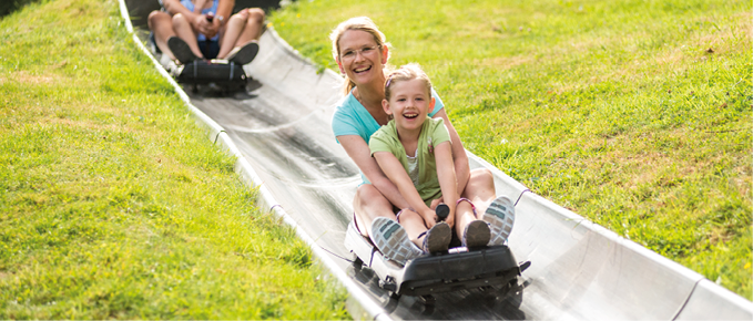 Das Bild zeigt eine Sommerrodelbahn im Freien auf einer gr nen Wiese.   Im Vordergrund sitzen eine Frau und ein Kind gemeinsam auf einem Schlitten. Das Kind sitzt vorne, die Frau dahinter. Beide lachen und schauen nach vorne, wirken fr hlich und aufgeregt. Das Kind h lt sich an den Griffen des Schlittens fest, w hrend die Frau es von hinten sichert.    Die Bahn besteht aus einer metallenen Rinne, die leicht absch ssig durch die Wiese verl uft.    Im Hintergrund folgen weitere Personen auf Schlitten, die ebenfalls die Bahn hinunterfahren.    Die Umgebung ist hell, sonnig und gr n, was auf einen warmen Tag im Freien hindeutet.