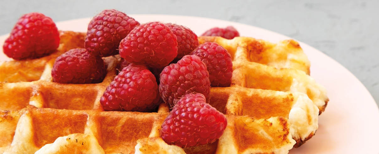 Das Bild zeigt eine goldbraun gebackene Waffel auf einem wei en Teller in Nahaufnahme.   Auf der Waffel liegen mehrere frische, rote Himbeeren, die sich in der Mitte zu einem kleinen Haufen stapeln. Die Oberfl che der Waffel ist gleichm  ig gerastert und wirkt au en knusprig, innen weich.    Die Himbeeren sehen saftig und leicht gl nzend aus und bilden einen farblichen Kontrast zur warmen, goldenen Waffel.    Der Hintergrund ist unscharf und hell, sodass der Fokus ganz auf der Waffel und den Fr chten liegt.