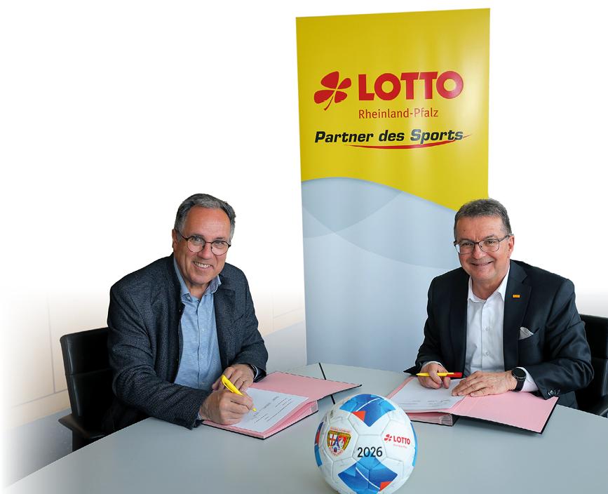 Zwei M nner in Anz gen sitzen an einem Besprechungstisch. Vor ihnen auf dem Tisch liegen ge ffnete Unterschriftenmappen. Diese sind aufgeschlagen und es hat den Anschein, dass die beiden Herren Unterlagen unterschreiben. Mitten auf dem Tisch liegt ein blau-wei er Fu ball mit den Logos des Fu ballverbands Rheinland sowie von Lotto Rheinland-Pfalz. Hinter den beiden steht ein Roll-Up. Darauf steht: “Lotto Rheinland-Pfalz - Partner des Sports".