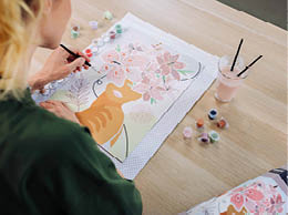 Person malt mit Pinsel ein Bild einer Katze und Blumen auf Papier mit mehreren kleinen Farbt pfchen und einem Getr nk auf dem Tisch