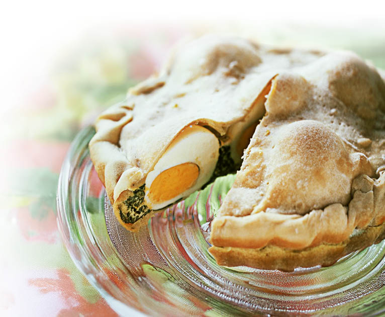 Ein runder, goldbraun gebackener Kuchen ist angeschnitten auf einer Glasplatte und zeigt im Inneren mehrere harte Eier, die halbiert eingebacken sind. Die F llung um die Eier wirkt gr nlich, vermutlich Spinat oder Kr uter, und bildet eine deutliche Schicht unter der Teigh lle.