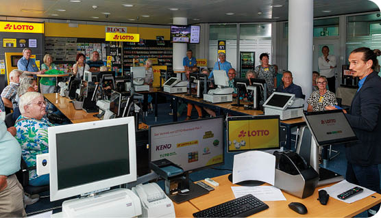 Auf dem Bild ist ein Schulungs- oder Informationsraum von LOTTO Rheinland-Pfalz zu sehen. Mehrere Personen sitzen oder stehen zwischen Verkaufstheken und Kassenarbeitspl tzen, die mit Monitoren, Druckern und Terminals ausgestattet sind.   Im Vordergrund steht ein Mann im Anzug, der offenbar einen Vortrag h lt oder eine Einweisung gibt. Die Anwesenden –  berwiegend  ltere Personen – h ren aufmerksam zu. An den Theken sind Logos und Hinweise zu LOTTO, KENO und weiteren Produkten angebracht.    Die Umgebung  hnelt einem realit tsnah eingerichteten Verkaufsraum, wie er in einer Lotto-Annahmestelle zu finden ist.