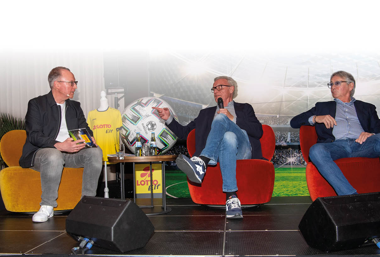 Das Bild zeigt eine Talkrunde auf einer B hne mit drei M nnern.   Die M nner sitzen jeweils in Sesseln – links auf einem gelben, in der Mitte und rechts auf roten Sesseln. Sie sind locker gekleidet, tragen Sakkos, Hemden und Jeans.    Der Mann in der Mitte h lt ein Mikrofon in der Hand und spricht, w hrend er mit dem Finger auf den Mann links zeigt. Dieser h rt aufmerksam zu und h lt ein kleines Heft oder Karten in der Hand. Der Mann rechts sitzt leicht zur ckgelehnt und blickt zur Mitte.    Zwischen ihnen steht ein kleiner runder Tisch mit Getr nken. Daneben ist ein gro er Fu ball als Dekoration zu sehen sowie ein gelbes Trikot mit der Aufschrift „LOTTO“.    Der Hintergrund zeigt ein Stadionmotiv, wodurch die Veranstaltung thematisch dem Sport – insbesondere Fu ball – zugeordnet werden kann.