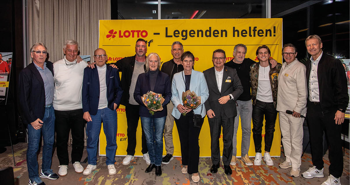 Das Bild zeigt eine Gruppe von etwa zw lf Personen, die nebeneinander f r ein Foto posieren.   Sie stehen vor einer gro en gelben Wand oder einem Banner mit dem roten Logo „LOTTO“ und dem Schriftzug „Legenden helfen!“. Die Personen sind  berwiegend erwachsen bis  lter, gemischt aus M nnern und Frauen, und tragen eine Mischung aus freizeitlicher und eleganter Kleidung – darunter Sakkos, Hemden, Pullover und Jeans.    In der Mitte stehen zwei Frauen, die jeweils einen Blumenstrau  in den H nden halten. Die  brigen Personen stehen dicht daneben, einige haben den Arm freundschaftlich um die Schultern der Nachbarn gelegt. Die Gruppe schaut in die Kamera und l chelt.