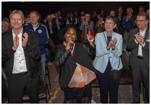 Das Bild zeigt eine lebendige Veranstaltung in einem Innenraum mit vielen stehenden Menschen.   Im Vordergrund steht eine Frau mit dunklen, lockigen Haaren, die ein Mikrofon in der Hand h lt. Sie tr gt ein dunkles Outfit und l chelt strahlend. Mit der anderen Hand hebt sie einen Finger nach oben, als w rde sie das Publikum animieren oder einen Moment betonen.    Um sie herum stehen mehrere Personen, darunter M nner und Frauen, die klatschen und l cheln. Einige tragen Sakkos oder Jacken, andere eher sportliche Kleidung. Die Stimmung wirkt gel st und begeistert.    Im Hintergrund sind weitere Menschen zu sehen, ebenfalls stehend und applaudierend. Die Beleuchtung ist ged mpft, typisch f r eine Veranstaltung oder ein Event.