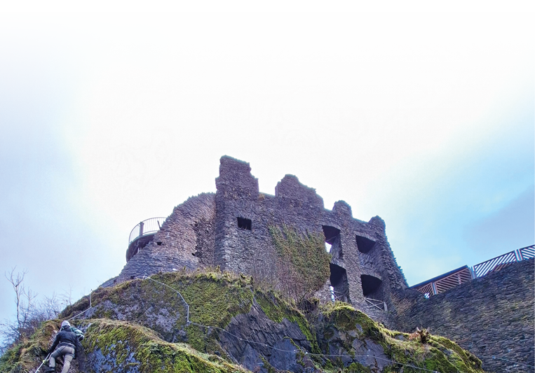 Das Bild zeigt die Ruine einer alten Burg auf einem felsigen H gel.   Die Mauerreste bestehen aus grauem Stein und wirken teilweise zerfallen. Mehrere Fenster ffnungen ohne Glas sind sichtbar, ebenso unregelm  ige Mauerkanten und eingest rzte Bereiche. Die Struktur ist kantig und massiv.    Die Burg steht auf einem steilen Felsen, der mit Moos und vereinzelter Vegetation bewachsen ist. Ein schmaler Weg oder Zugang f hrt seitlich nach oben.    Der Himmel im Hintergrund ist hell und leicht bew lkt, wodurch die Ruine etwas d ster und zugleich eindrucksvoll wirkt.