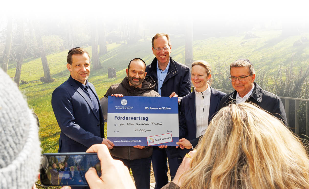 Das Bild zeigt eine Gruppe von f nf Personen, die im Freien zusammenstehen und f r ein Foto posieren. Die Gruppe h lt gemeinsam eine gro e F rderurkunde bzw. einen symbolischen Scheck mit der  berschrift „F rdervertrag“. Darauf ist ein Betrag von 95.000 Euro angegeben sowie ein Hinweis auf die Deutsche Stiftung Denkmalschutz und die F rderinitiative Gl cksSpirale. Der F rderzweck bezieht sich auf einen j dischen Friedhof.  Die Personen – drei M nner und zwei Frauen – sind halb formell gekleidet und l cheln in die Kamera. Sie stehen eng beieinander und halten das Dokument gut sichtbar nach vorne. Im Vordergrund ist eine weitere Person zu sehen, die mit einem Smartphone ein Foto der Gruppe macht. Das Smartphone-Display zeigt die gleiche Szene.  