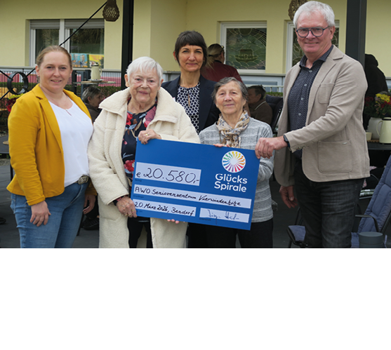 Das Bild zeigt f nf Personen unterschiedlichen Alters, die gemeinsam im Freien f r ein Foto posieren. Im Vordergrund halten zwei  ltere Frauen einen gro en blauen Spendenscheck. Der Scheck tr gt das Logo „Gl cksSpirale“ und weist einen Betrag von 20.580 Euro aus. Als Empf nger ist ein Seniorenzentrum angegeben.  Links und rechts neben den beiden Frauen stehen drei weitere Personen: eine Frau in einem gelben Blazer, eine Frau in dunkler Kleidung sowie ein Mann mit Brille und hellem Sakko. Alle blicken in die Kamera und l cheln.  Die Gruppe steht vor einem Geb ude mit Fenstern und Terrasse, im Hintergrund sind weitere Menschen unscharf zu erkennen.  