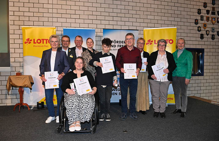 Das Bild zeigt eine Gruppe von elf Erwachsenen bei einer feierlichen Veranstaltung in einem Innenraum. Die Personen sind in zwei Reihen angeordnet, wobei in der vorderen Reihe eine Frau im Rollstuhl mittig sitzt und von mehreren stehenden Personen flankiert wird, w hrend dahinter weitere M nner und Frauen dicht nebeneinander stehen. Alle wirken wie Teil einer gemeinsamen Ehrung oder Auszeichnung. Die meisten halten Urkunden mit der berschrift „Ehrenurkunde“ gut sichtbar vor sich. Die Gruppe besteht  berwiegend aus Personen mittleren bis h heren Alters, die gepflegte, halb-formelle Kleidung tragen, darunter Blazer, Hemden, Blusen und Pullover in eher gedeckten Farben wie Schwarz, Blau, Grau und Beige. Einige tragen Brillen, und die meisten blicken in die Kamera mit freundlichem oder leicht l chelndem Gesichtsausdruck.    Die Frau im Rollstuhl im Vordergrund tr gt ein schwarzes Oberteil und einen Rock mit auff lligem schwarz-wei em Muster. Sie sitzt aufrecht und h lt ihre Urkunde mit beiden H nden auf dem Scho , ihr Ausdruck wirkt ruhig und zufrieden. Die  brigen Personen stehen eng beieinander und pr sentieren ebenfalls ihre Urkunden, was den gemeinschaftlichen Charakter der Ehrung unterstreicht.    Im Hintergrund befindet sich eine helle, wei  gekachelte Wand. Dahinter stehen mehrere gro e Roll-up-Banner, darunter gelbe Banner mit der Aufschrift „LOTTO Rheinland-Pfalz“ sowie blaue Banner mit teilweise lesbaren Worten wie „F rder…“ und „Fre…“, die auf eine Organisation oder F rderinitiative hinweisen. Rechts an der Wand sind zahlreiche kleine Wappen oder Abzeichen angebracht. Auf der linken Seite des Bildes steht ein kleiner runder Tisch mit einem goldfarbenen Tuch, auf dem ein kleines Objekt liegt, vermutlich eine Auszeichnung oder Dekoration.    Die Beleuchtung ist gleichm  ig und hell, ohne starke Schatten, und der Boden wirkt dunkel, m glicherweise ein Teppich oder B hnenbelag. Insgesamt vermittelt das Bild eine offizielle, respektvolle und positive Atmosph re, wie sie typisch f r eine Preisverleihung oder eine  ffentliche Ehrung ist.