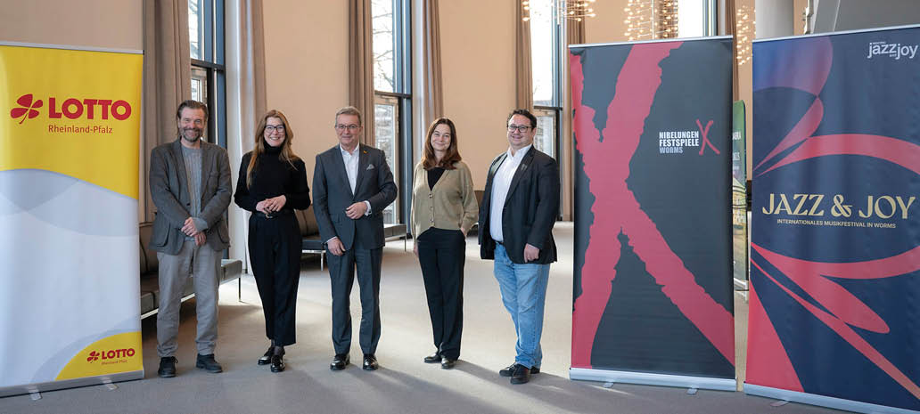 Das Bild zeigt f nf Personen, die nebeneinander in einem hellen Innenraum stehen und in die Kamera l cheln. Die Gruppe besteht aus drei M nnern und zwei Frauen, die  berwiegend gesch ftlich bis elegant gekleidet sind – mit Sakkos, Blazern, Hemden und Stoffhosen.  Links im Bild steht ein gelbes Banner mit dem Logo „LOTTO Rheinland-Pfalz“. Rechts daneben befinden sich zwei weitere Roll-up-Banner: eines mit der Aufschrift „Nibelungen-Festspiele Worms“ und ein weiteres mit „Jazz & Joy – Internationales Musikfestival in Worms“.  Der Raum wirkt gro z gig und modern, mit hohen Fenstern, durch die Tageslicht einf llt. Im Hintergrund sind Sitzgelegenheiten entlang der Wand zu erkennen.  Die Personen stehen entspannt mit leichtem Abstand zueinander und haben eine offene, freundliche Haltung.  