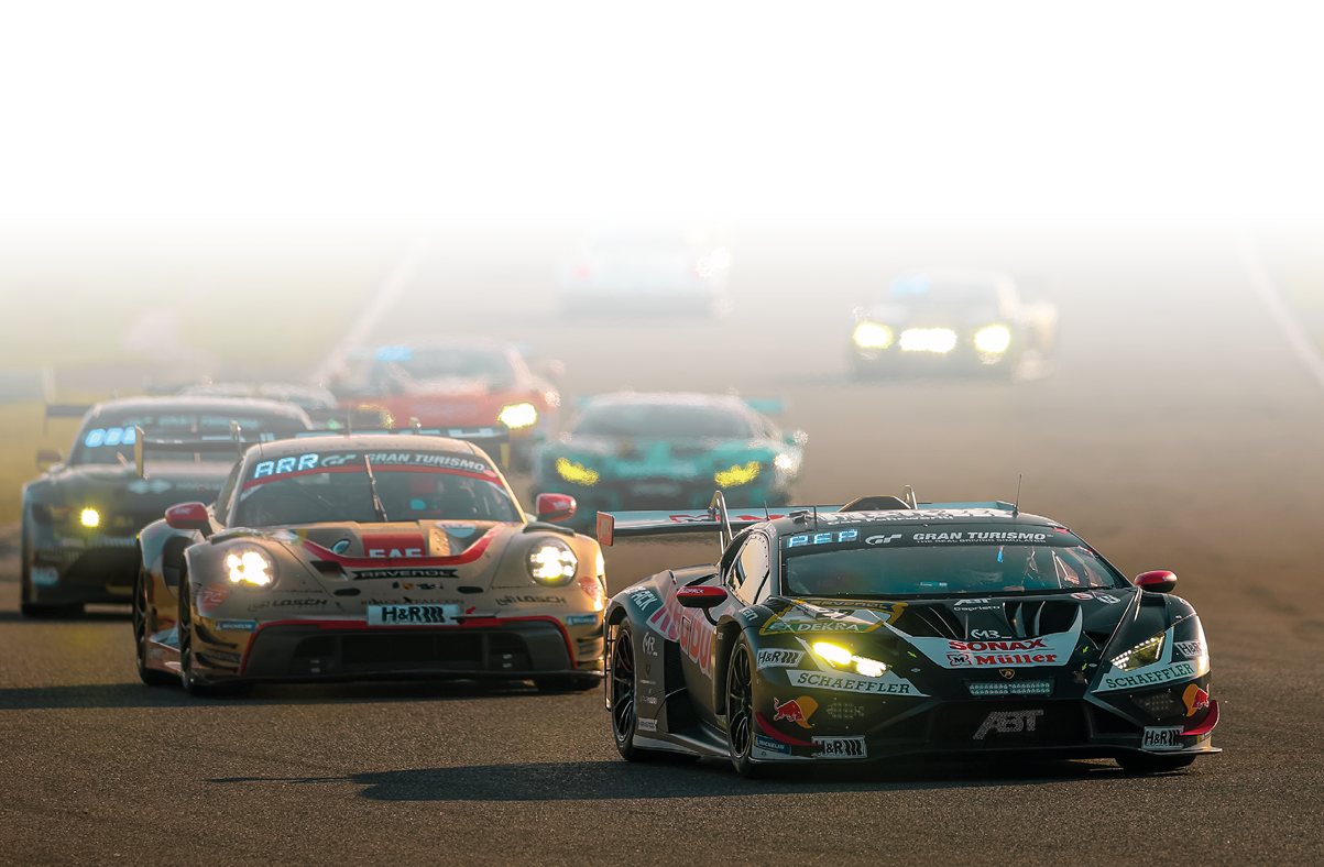 Im Vordergrund fahren mehrere flache, breite Rennwagen (GT-Sportwagen) dicht hintereinander auf einer asphaltierten Strecke. Das f hrende Fahrzeug ist schwarz mit gr nen und roten Sponsorenaufklebern und hat eingeschaltete Scheinwerfer. Direkt dahinter folgt ein heller Rennwagen mit roten Akzenten. Weitere Fahrzeuge sind im Hintergrund zu sehen, teilweise unscharf, was die hohe Geschwindigkeit und Bewegung unterstreicht.    Das Licht ist warm, vermutlich sp ter Nachmittag oder Abend, wodurch die Szene in goldene Farbt ne getaucht ist.      