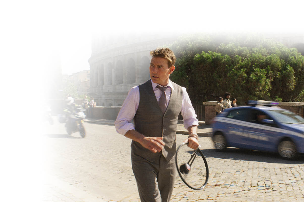 Das Bild zeigt einen Mann in Bewegung auf einer Stra e in einer historischen Stadt.   Er tr gt ein helles Hemd, eine Weste und eine Krawatte und wirkt elegant gekleidet. In einer Hand h lt er Kopfh rer, w hrend er sich z gig nach vorne bewegt. Sein Blick ist nach rechts gerichtet, als w rde er auf etwas reagieren oder sich orientieren.    Im Hintergrund ist deutlich das Kolosseum in Rom zu erkennen. Die Umgebung zeigt eine st dtische Szene mit Kopfsteinpflaster, vorbeifahrenden Fahrzeugen und einigen Menschen am Stra enrand.    Rechts f hrt ein blaues Polizeiauto vorbei, dessen Bewegung leicht verschwommen ist, was Dynamik vermittelt. Auch ein Motorrad ist im Hintergrund zu sehen.    Die linke Bildseite ist stark aufgehellt bzw. ausgeblendet, wodurch der Fokus auf dem Mann und der Szene rechts liegt.