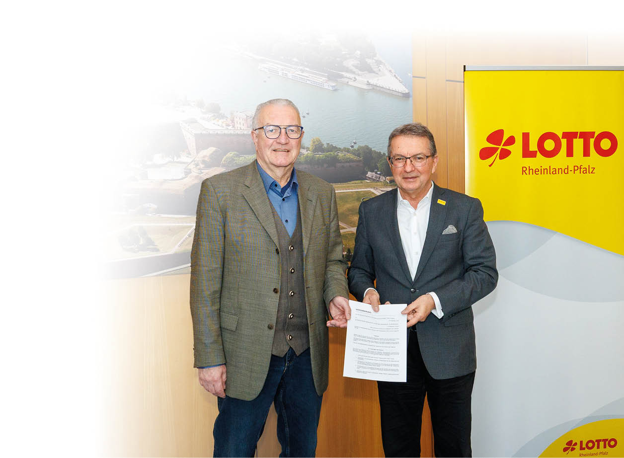 Das Bild zeigt zwei M nner, die nebeneinander in einem Innenraum stehen und gemeinsam ein Dokument in den H nden halten. Beide sind gesch ftlich gekleidet: Der Mann links tr gt ein Sakko mit Weste und Hemd, der Mann rechts einen dunklen Anzug mit Hemd. Sie schauen in die Kamera und wirken ruhig und freundlich.  Hinter ihnen ist ein gro es Wandbild zu sehen, das eine Luftaufnahme der Stadt Koblenz mit Fluss und Festungsanlage zeigt. Rechts daneben steht ein gelbes Roll-up-Banner mit dem Logo „LOTTO Rheinland-Pfalz“.  Die Umgebung wirkt wie ein B ro oder Konferenzraum mit Holzverkleidung an den W nden.  