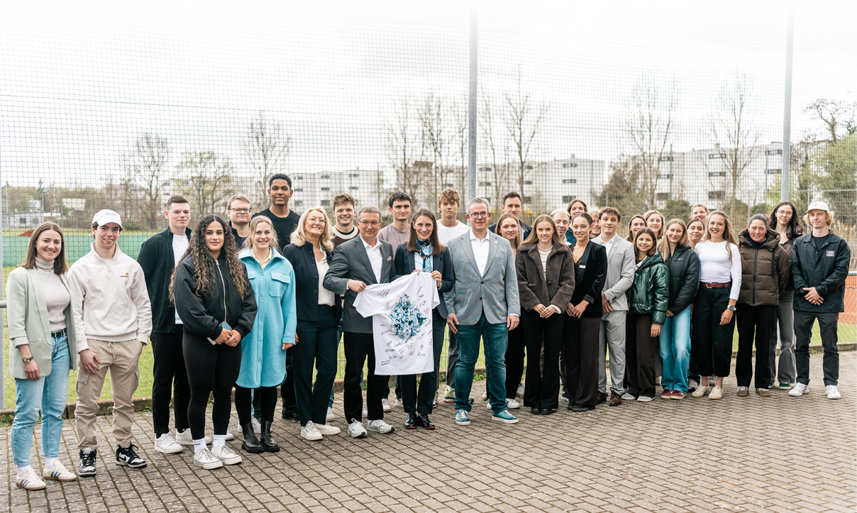 Das Bild zeigt eine gro e Gruppe von rund 25 bis 30 Personen, die nebeneinander im Freien f r ein Foto posieren. Die Gruppe besteht  berwiegend aus jungen Erwachsenen, erg nzt durch einige  ltere Personen. Sie stehen auf einem gepflasterten Platz vor einem Sportgel nde, erkennbar an einem Zaun und Spielfeld im Hintergrund.  In der Mitte halten zwei Personen ein wei es T-Shirt, das mit Unterschriften und einem blauen Motiv versehen ist.  Die Kleidung ist locker bis halb formal, von Jeans und Sneakern bis hin zu Blazern und M nteln. Die meisten blicken in die Kamera und l cheln.  Im Hintergrund sind Geb ude und B ume zu sehen, die auf eine st dtische Umgebung hindeuten.