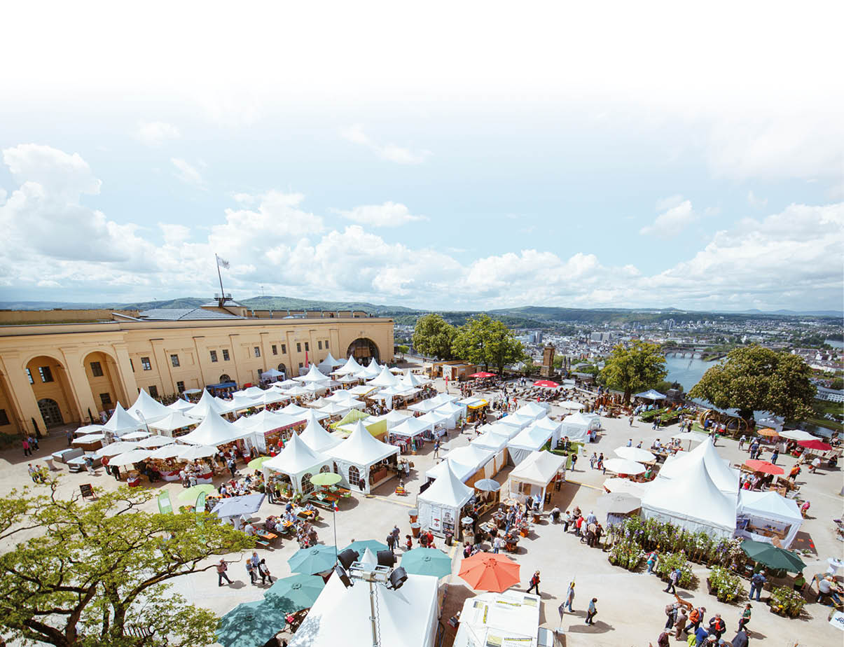 Das Bild zeigt einen gro en, lebhaften Markt aus der Vogelperspektive.   Auf einem weitl ufigen Platz stehen zahlreiche wei e Zelte und Marktst nde, die in Reihen angeordnet sind. Dazwischen bewegen sich viele Menschen, die bummeln, einkaufen oder an Tischen sitzen.    Links im Bild befindet sich ein gro es, helles Geb ude mit klassischer Architektur, es ist ein Teil der Festung Ehrenbreitstein in Koblenz. Rund um den Platz stehen au erdem B ume, die Schatten spenden.    Zwischen den Zelten sind bunte Sonnenschirme zu sehen, unter denen Besucher sitzen oder sich ausruhen. Einige St nde bieten Pflanzen, Kunsthandwerk oder Speisen an.    Im Hintergrund  ffnet sich der Blick auf Koblenz, die Mosel und Br cken, eingebettet in eine leicht h gelige Landschaft. Der Himmel ist blau mit lockeren wei en Wolken, was auf einen sonnigen Tag hindeutet.