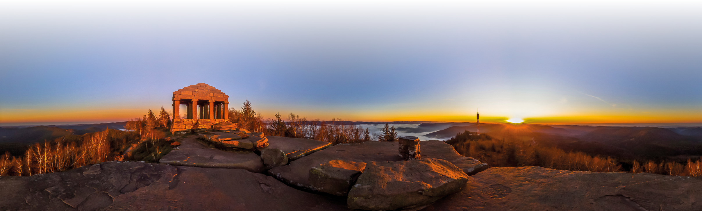 Das Bild zeigt eine weite Panorama‑Landschaft bei Sonnenuntergang oder Sonnenaufgang. Im Vordergrund liegt eine gro e, flache Felsplatte mit Rissen und eingravierten Zeichen, die wie ein Aussichtsfelsen wirkt. Links steht ein kleiner, rundlicher Steinpavillon mit S ulen, wie ein Denkmal. In der Mitte und rechts sieht man W lder und H gel, die sich bis zum Horizont. Ganz rechts am Horizont steht die Sonne knapp ber der Landschaft; der Himmel geht von dunklem Blau oben zu orangegelb am Horizont  ber. Die Szene wirkt ruhig und weit, ohne sichtbare Personen.