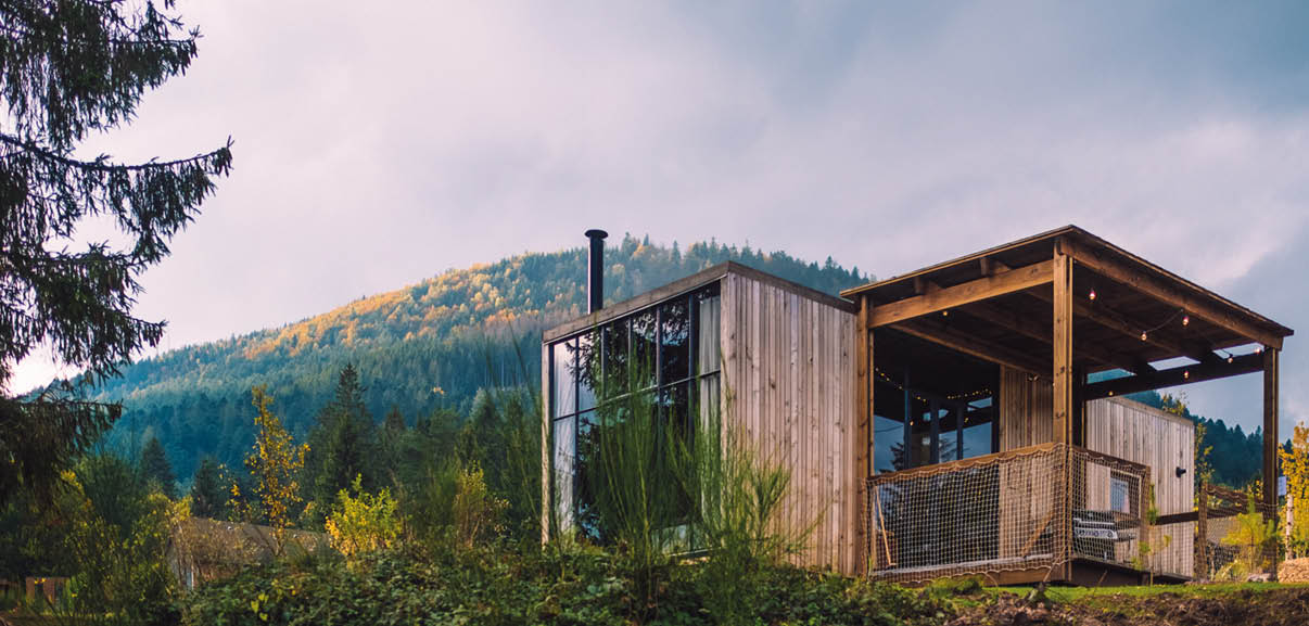 Das Bild zeigt ein modernes, kleines Holzhaus oder eine H tte am Rand eines Waldes, mit Blick auf bewaldete H gel im Hintergrund. Links am Geb ude ist eine gro e Glasfront, in der sich B ume spiegeln; auf dem Dach ragt ein schwarzes Ofenrohr nach oben. Rechts hat die H tte eine berdachte Terrasse mit Holzpfosten und einer Lichterkette unter dem Dach; an der Vorderkante h ngt ein Netz oder Gel nder. Links ragt der dunkle Stamm und  ste einer Tanne ins Bild. Der Himmel ist grau und wolkig, das Licht wirkt wie am sp ten Nachmittag oder Abend. Personen sind nicht zu sehen; die Szene wirkt ruhig und abgelegen.