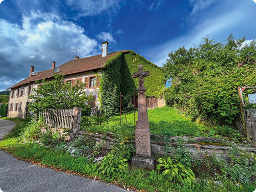 Zu sehen ist ein zweist ckiges, lteres Wohnhaus mit braunem Satteldach und hellen Au enw nden, fotografiert an einer schmale Dorfstra e. Die rechte Hausseite ist dicht mit W lder Efeu  berwachsen, w hrend links Fenster mit braunen Klappl den zu erkennen sind. Vor dem Haus liegt ein gr ner, etwas Schweizer Garten, eingefasst von einer niedrigen Steinmauer und einem schiefen Holzzaun. Im Vordergrund steht ein hohes steinernes Kreuz mit Christusfigur auf einem Sockel. Rechts am Bildrand ist ein Stra enschild nur teilweise sichtbar, der Text beginnt mit „de la…“ und ist sonst nicht lesbar.  ber allem spannt sich ein kr ftig blauer Himmel mit gro en wei en Wolken; die Szene wirkt sonnig und l ndlich, ohne sichtbare Personen.