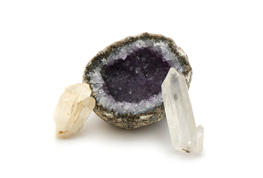 Auf wei em Hintergrund liegt in der Mitte ein aufgeschnittener Ametyst mit einer dunkellila, kristalligen Innenfl che. Der Rand der Geode ist grau bis br unlich und wirkt rau wie Stein. Links daneben liegt ein kleiner, milchig‑heller Kristallbrocken mit mehreren kantigen Spitzen. Rechts lehnt ein l nglicher, transparenter Kristall mit prismatischer Form an der Geode. Das Bild wirkt wie eine sachliche Produktaufnahme von Mineralien, ohne weitere Objekte oder Text.