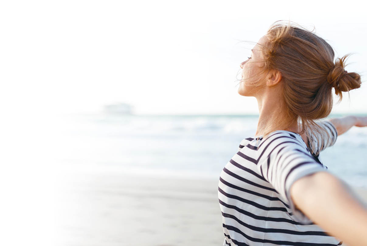 Am Strand steht eine junge Frau im Profil und blickt nach rechts Richtung Meer. Sie tr gt ein wei ‑dunkelblau gestreiftes Shirt und hat die Haare zu einem lockeren Knoten hochgebunden; einzelne Str hnen wehen im Wind. Beide Arme sind weit ausgebreitet, als w rde sie tief durchatmen oder die frische Luft genie en. Hinter ihr sieht man Sand, ruhige Wellen und einen hellen Himmel; am Horizont ist ein Objekt (m glicherweise ein Schiff oder Geb ude) unscharf. Die Stimmung wirkt ruhig, frei und entspannend.