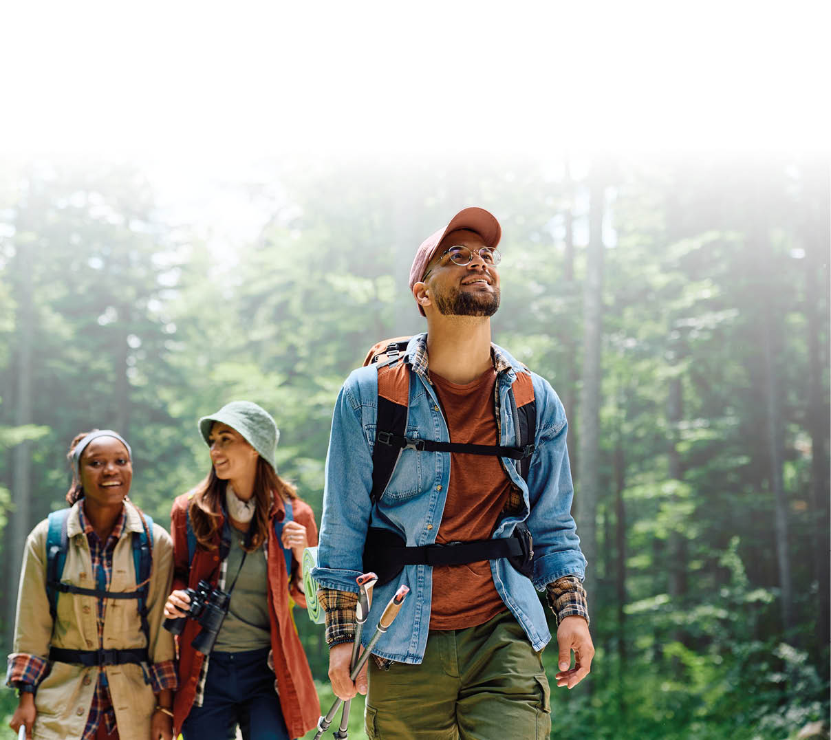 Im Vordergrund geht ein Mann mit Kappe, Brille und Rucksack auf einem Waldweg und blickt nach oben. Hinter ihm folgen zwei Frauen, ebenfalls mit Rucks cken. Eine tr gt einen Hut und h lt ein Fernglas, die andere l chelt. Alle drei wirken fr hlich und sind in Freizeit- bzw. Wanderkleidung gekleidet. Der Wald ist dicht bewachsen, gr n und von weichem Licht durchflutet, was eine ruhige, nat rliche Atmosph re vermittelt.