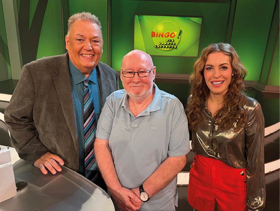Das Bild zeigt drei Personen in einem Fernsehstudio, die eng beieinanderstehen und in die Kamera l cheln. In der Mitte steht ein  lterer Mann mit Glatze und Brille, der ein hellblaues Poloshirt tr gt. Links neben ihm steht ein gr  erer Mann mit kurzen grauen Haaren, gekleidet in Sakko, Hemd und Krawatte. Rechts steht eine Frau mit langen, gewellten Haaren, die eine gl nzende Bluse und einen roten Rock tr gt.  Alle drei wirken freundlich und entspannt.  Im Hintergrund ist ein gr n beleuchtetes Studio zu sehen. Auf einem Bildschirm steht deutlich der Schriftzug „BINGO!“, was auf eine Spiel- oder Unterhaltungssendung hinweist.  Vorne links ist ein Teil eines Tisches oder Pults sichtbar.  