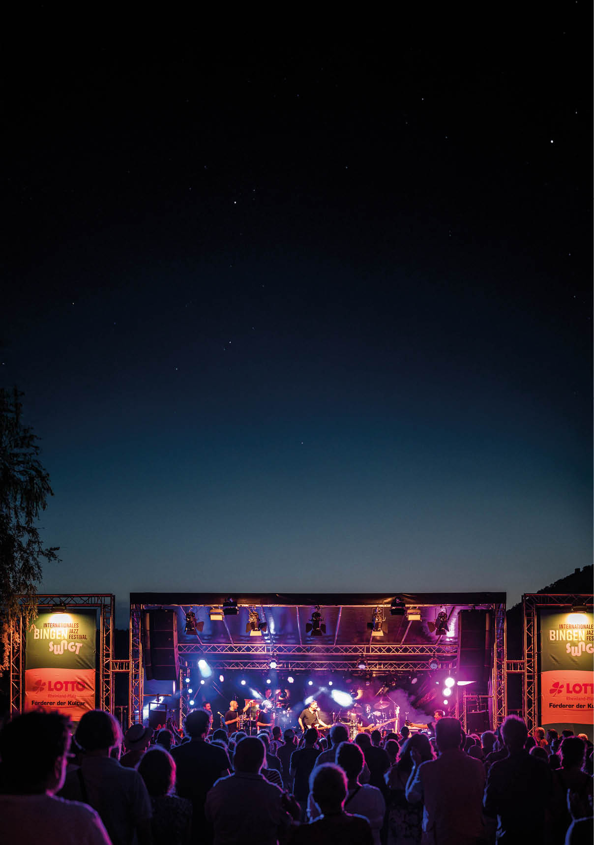 Panoramaaufnahme einer Open-Air-Konzertb hne bei Nacht. Im Vordergrund sind viele Zuschauerinnen und Zuschauer als dunkle Silhouetten zu sehen, dicht gedr ngt und auf die B hne gerichtet. Auf der B hne spielt eine gr ere Band mit mehreren Musikerinnen und Musikern; in der Mitte steht eine Person im Rampenlicht. Kr ftige B hnenbeleuchtung in Blau-, Violett- und Wei t nen strahlt nach vorn, Rauch- oder Nebeleffekte machen die Lichtkegel sichtbar. Links und rechts der B hne h ngen hohe Banner mit der Aufschrift „Internationales Bingen Swingt Jazz Festival“; darunter ist ein Sponsorenhinweis „LOTTO Rheinland-Pfalz – F rderer der Kultur“ zu erkennen.