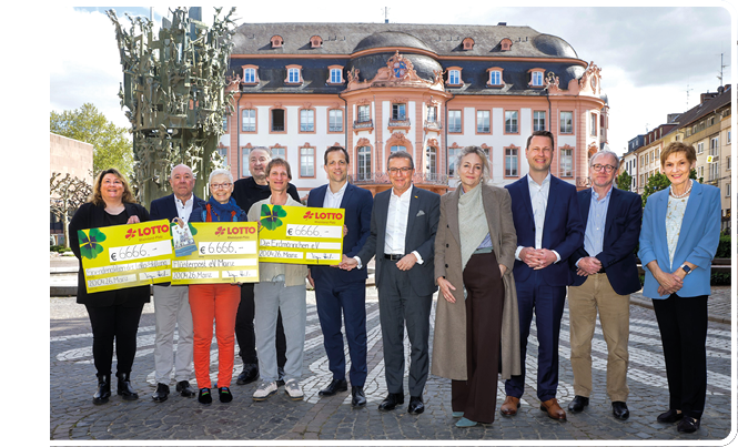 Auf dem Foto steht eine Gruppe von zw lf Erwachsenen nebeneinander auf einem hellen, rund gepflasterten Platz. Im Hintergrund ist ein mehrst ckiges, rosa-wei es Barockgeb ude mit geschwungenem Dach sowie Fenstern und Stuckverzierungen zu sehen; links im Bild steht eine hohe, abstrakte bronzene Skulptur. Die Gruppe ist mittig im Bild angeordnet, alle blicken in Richtung Kamera. Die drei sichtbaren gelben Schecks sind gro formatig und gut lesbar; auf jedem Scheck steht prominent „LOTTO“ sowie der Betrag „€ 6.666,-“. Auf einem Scheck ist zus tzlich handschriftlich ein Empf ngername. Die Stimmung wirkt formell und feierlich, wie bei einer Scheck bergabe oder Spenden bergabe im  ffentlichen Raum. Das Tageslicht ist hell, der Himmel leicht bew lkt. Keine der abgebildeten Personen ist einzeln hervorgehoben; das Bild vermittelt Gemeinschaft und  ffentlichkeitsarbeit.