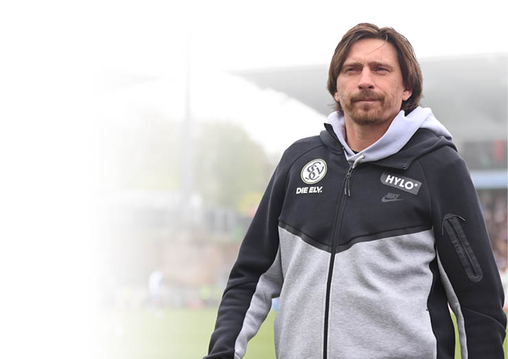 Ein Mann steht am Rand eines Fu ballfeldes und blickt konzentriert nach vorne. Er tr gt eine schwarz-graue Sportjacke mit Logos und hat mittellanges braunes Haar sowie leichten Bart. Im Hintergrund ist ein Stadion mit Zuschauern und Spielern zu sehen, die unscharf dargestellt sind. Die Szene wirkt wie ein Moment w hrend eines Spiels oder Trainings.