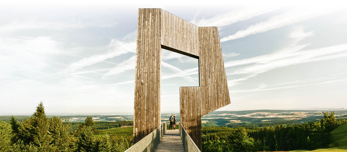 Das Foto zeigt eine erh hte Aussichtsplattform aus Holz. Ein gerader Holzsteg f hrt mittig nach vorn und endet in einer gro en, senkrecht stehenden Holzkonstruktion, die wie ein berdimensionaler, kantiger Rahmen mit rechteckiger  ffnung wirkt. Am Ende des Stegs stehen zwei Menschen und schauen in die Landschaft. Links und rechts unterhalb der Plattform erstreckt sich ein dichtes Waldgebiet; dahinter sind Felder und sanfte H henz ge zu erkennen.  ber der Szene liegt ein heller Himmel mit d nnen, streifigen Wolken.