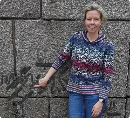 Zu sehen ist eine l chelnde Frau, die vor einer gro en, grauen Steinwand steht. In die Wand ist eine Reliefdarstellung eingearbeitet, die wie eine Karte beziehungsweise ein Flusslauf wirkt. Die Frau tr gt einen blau gemusterten Pullover und Jeans und zeigt mit der linken Hand auf einen markierten Punkt in der Reliefdarstellung. 
