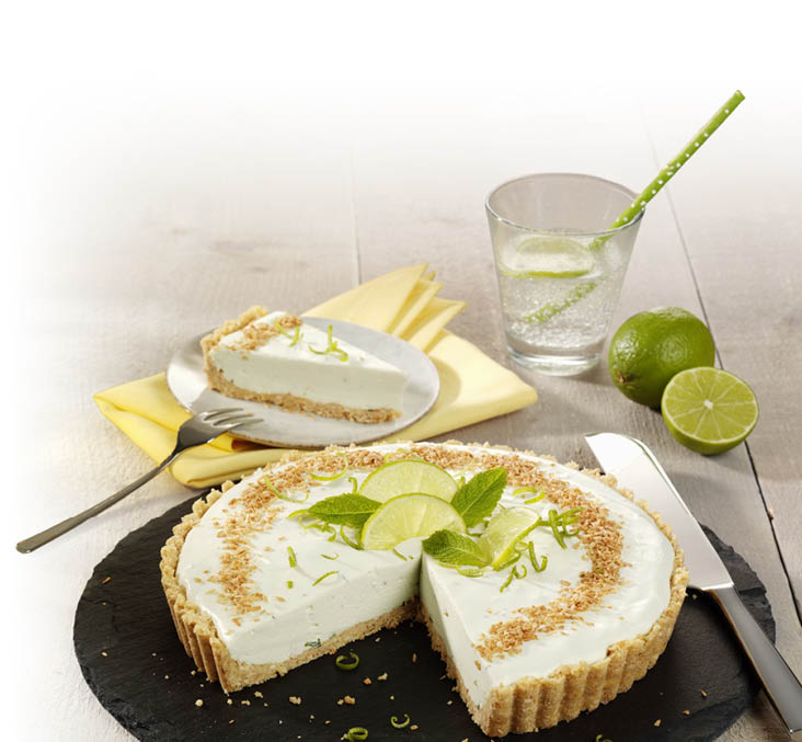 Auf dem Bild sieht man eine runde Torte mit heller Cremef llung und einem hellen Keksboden auf einer schwarzen Servierplatte. Aus der Torte ist ein gro es St ck herausgeschnitten; das St ck liegt hinten auf einem kleinen Teller auf einer gelben Serviette. Oben auf der Torte liegen Limettenscheiben, Limettenschale und gr ne Minzbl tter; am Rand sind ger stete Kr mel gestreut. Rechts steht ein Glas mit sprudelndem Getr nk, darin ist eine Limettenscheibe und ein gr ner Strohhalm; daneben liegen eine ganze Limette und eine halbe Limette. Vorne rechts liegt ein Messer, links eine Gabel auf dem Holztisch.