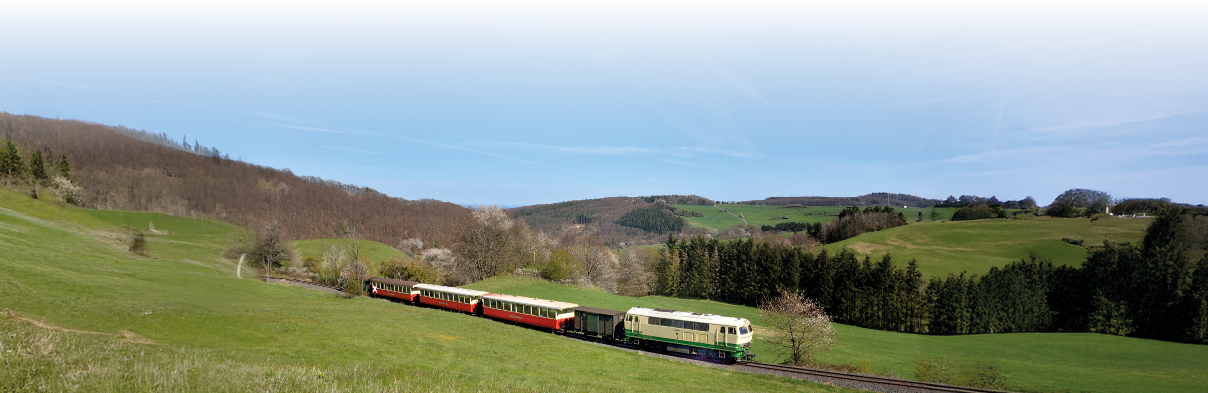 Im Vordergrund verl uft eine weite, sanft abfallende Wiese in sattem Fr hlingsgr n. Auf einer leicht erh hten Gleislinie f hrt von rechts nach links ein schmalspuriger Dieselzug. Die Lokomotive ist hellgr n bis cremefarben, direkt dahinter liegt ein kleiner offener G terwagen, gefolgt von zwei kurzen, roten Personenwagen mit hellen Dachpartien. Links und rechts der Gleise stehen vereinzelt bl hende B ume und Str ucher; im linken Bilddrittel schlie en sich bewaldete H nge mit kahlen bis leicht belaubten B umen an. Im Hintergrund ziehen sich sanfte, bewaldete H gel bis zum Horizont, darunter einzelne hellergr ne Lichtungen und Wiesen. Der Himmel ist klar und blau mit wenigen, feinen Kondensstreifen. Die Szene wirkt ruhig und l ndlich, typisch f r eine fr he Fr hlingstagefahrt durch eine Mittelgebirgslandschaft.