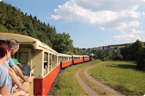 Das Bild zeigt einen l ngeren, rot‑beigen Museumszug, der von links vorn nach rechts hinten durch eine l ndliche Tallandschaft f hrt. Im linken Vordergrund steht das offene Ende eines Personenwagens; dort lehnen mehrere Fahrg ste mit Blick nach rechts aus dem Wagen heraus. Der Zug besteht aus mehreren kurz gekuppelten Wagen mit gro en rechteckigen Fenstern; einige Fenster sind ge ffnet, durch sie sind weitere Mitreisende und Details der Innenr ume zu erkennen.   Rechts neben den Gleisen erstreckt sich eine breite Wiese mit hohem Gras, durch die ein geschwungener, leicht ausgefahrener Feldweg f hrt, der dem Zugverlauf folgt. Links im Bild steigt der Hang mit dichten B umen und Waldrand auf; der Zug f hrt entlang dieser Waldkante. Im Hintergrund, rechts hinten, ist ein kleines Dorf sichtbar mit vereinzelten H usern und Geb uden, hinter denen weitere bewaldete H gel aufragen.    Der Himmel ist gro teils blau mit einigen wei en, flauschigen Wolken. Die Lichtstimmung ist hell und sonnig, typische Sommertags‑Atmosph re. Die Szene vermittelt ein Gef hl von Ausflug, Bewegung und l ndlicher Idylle.